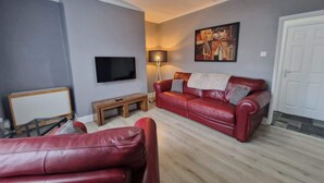 Ein Wohnzimmer mit einer roten Ledersofa, einer Holzbank, einem Flachbildfernseher, einer Stehlampe und einem Gemälde an der Wand.