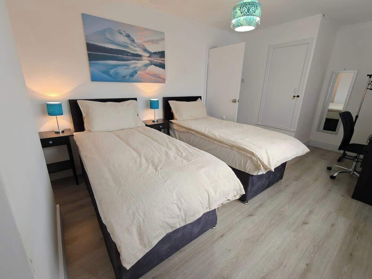 Quarto com duas camas, uma mesa de cabeceira com abajur, um quadro na parede e uma porta.