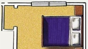 Una habitación con cama, una mesita, una silla, un ventanal y un baño con lavamanos e inodoro.