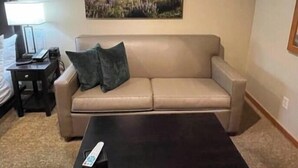 Una sala de estar con un sofá color beige, una mesa de centro oscura, un control remoto, una lámpara y un cuadro enmarcado en la pared.