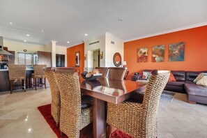 Ein Essbereich mit Holztisch und gewebten Stühlen, im Hintergrund eine Küche und eine orangefarbene Wand mit Kunstwerken.