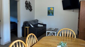 Een eetgedeelte met een houten tafel, stoelen, een aan de muur bevestigde televisie en een schilderij.