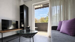 A modern living room with a flat-screen TV, a glass table, a sofa with purple cushions, and a sliding glass door leading to an outdoor area.