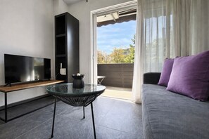 Een moderne woonkamer met een flatscreen televisie, een glazen tafel, een bank met paarse kussens en een schuifpui naar een buitenruimte.