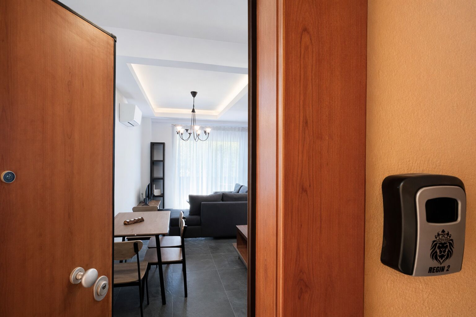Habitación de hotel con puerta de madera, una mesa con sillas, un sofá, una araña y un dispositivo montado en la pared con la etiqueta 'REGIN 2'.