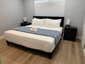 A neatly made bed with a white duvet and a blue blanket, a black nightstand with a lamp, and a wooden floor.