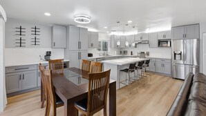 A modern kitchen with white cabinets, stainless steel appliances, a dining area with wooden chairs, and a leather sofa. - Modern 4BR Home, Sleeps 9, Near Silicon Valley (San Jose)