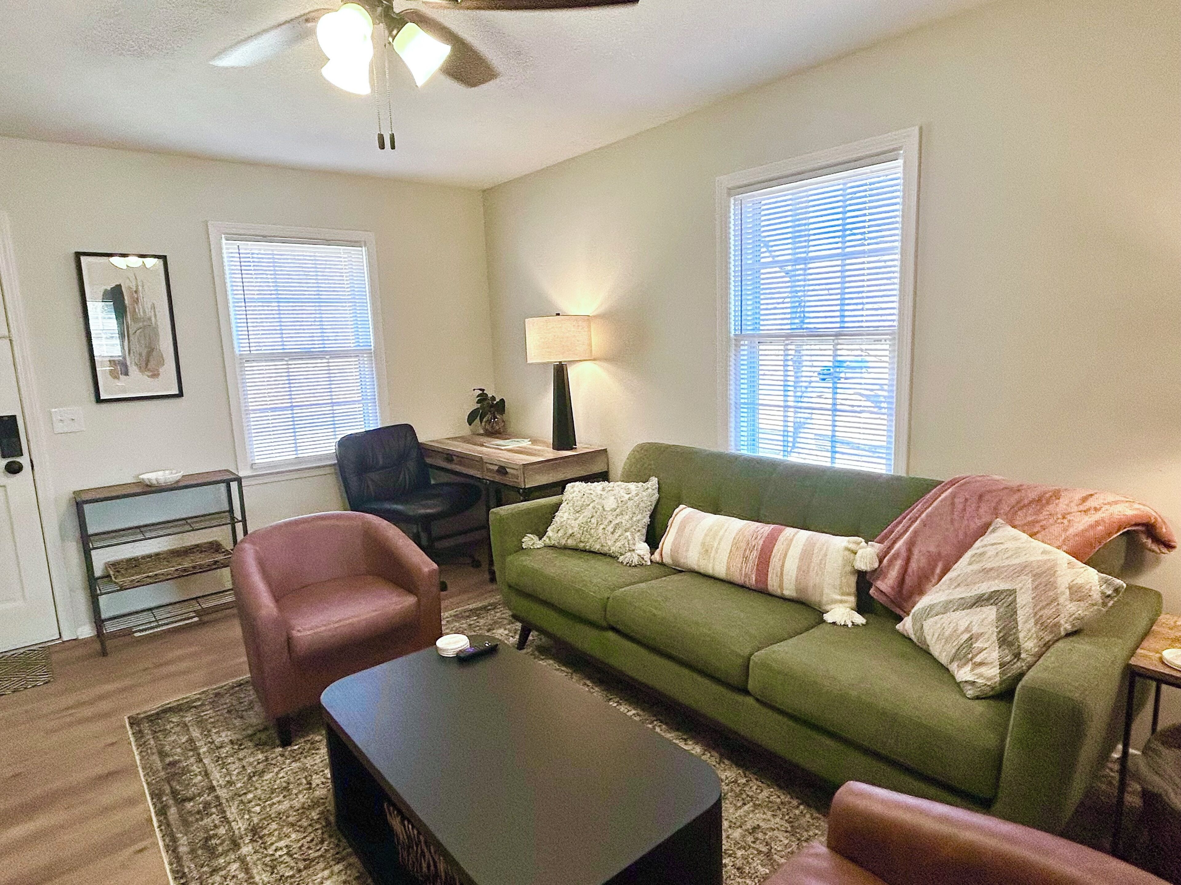 A living room with a green sofa, a black armchair, a coffee table, a desk with a lamp, and a ceiling fan.