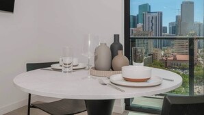 Uma mesa de jantar redonda com toalha branca, posta com copos transparentes, pratos e tigelas, e vasos em um quarto moderno com vista para a cidade.