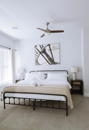 Une chambre à coucher comprenant un lit, un ventilateur de plafond, des œuvres d’art, des lampes de chevet et une fenêtre avec des stores.