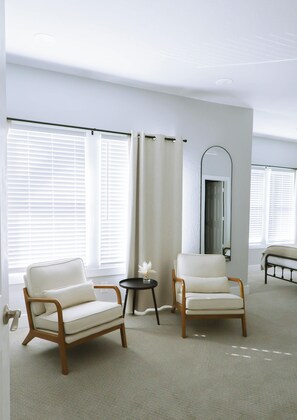Une chambre moderne avec deux fauteuils blancs, une petite table ronde ornée d’un vase et un lit avec une tête de lit.