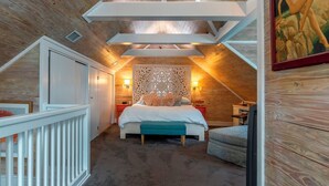 Un dormitorio con techo inclinado, cama, mesita de noche, silla y lámpara de pared.