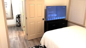 Et hotelværelse med seng, TV på et stativ, en dør og et vindue med persienner.