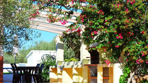 An outdoor patio with a grill, a table with chairs, and a pergola with flowers.