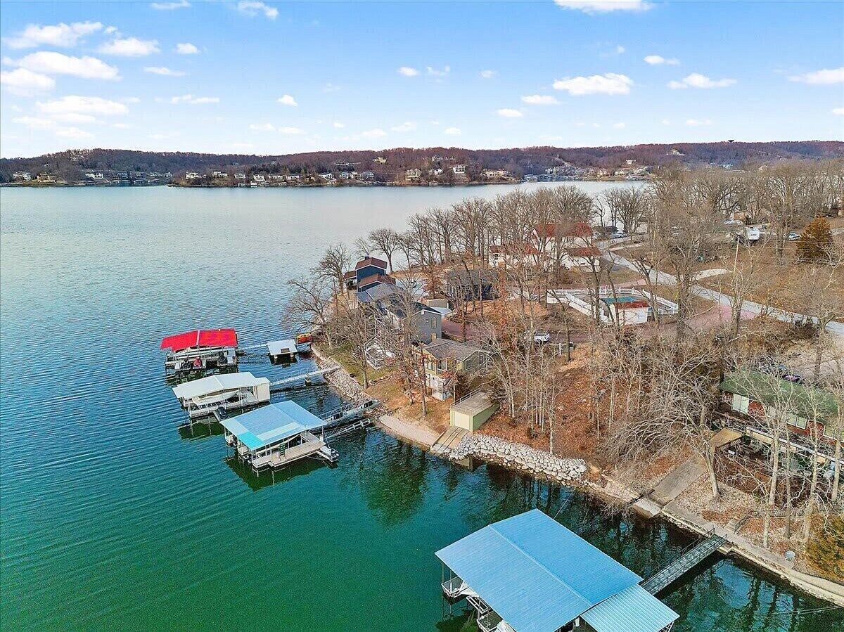 An aerial view of a lakeside property with docked boats, a stone bridge, and houses surrounded by trees.