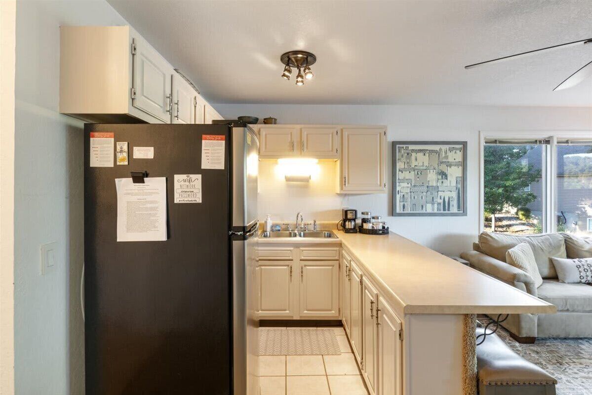 Una cocina moderna con armarios blancos, un refrigerador negro y un comedor con mesa y sofá de colores claros.