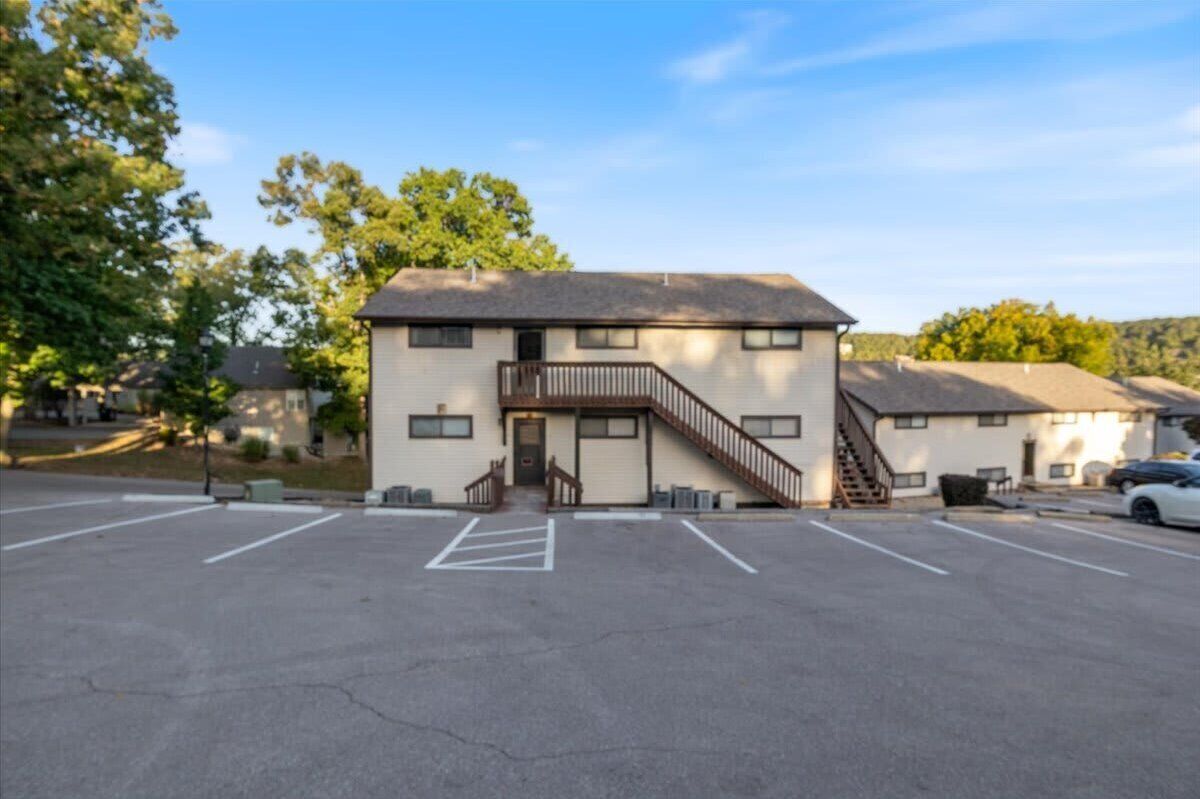 A two-story building with a staircase leading to the entrance, a parking lot in front, and trees in the background.