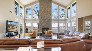 A spacious living room with a stone fireplace, leather sofas, and large windows offering a view of the outdoors. - Lakefront House, Dock, Fire pit (Sunrise Beach)