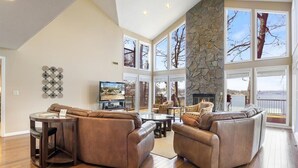 A spacious living room with a stone fireplace, large windows, and a high ceiling. - Lakefront House, Dock, Fire pit (Sunrise Beach)