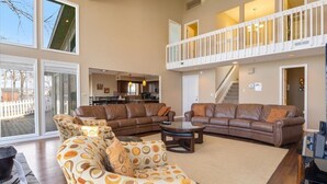 A spacious living room with a brown leather sofa set, a patterned armchair, and a coffee table. A staircase leads to an upper level with a balcony. - Lakefront House, Dock, Fire pit (Sunrise Beach)