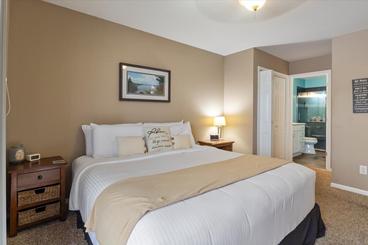 A bedroom with a large bed, a nightstand with a lamp, a framed picture on the wall, and a bathroom visible through an open door.