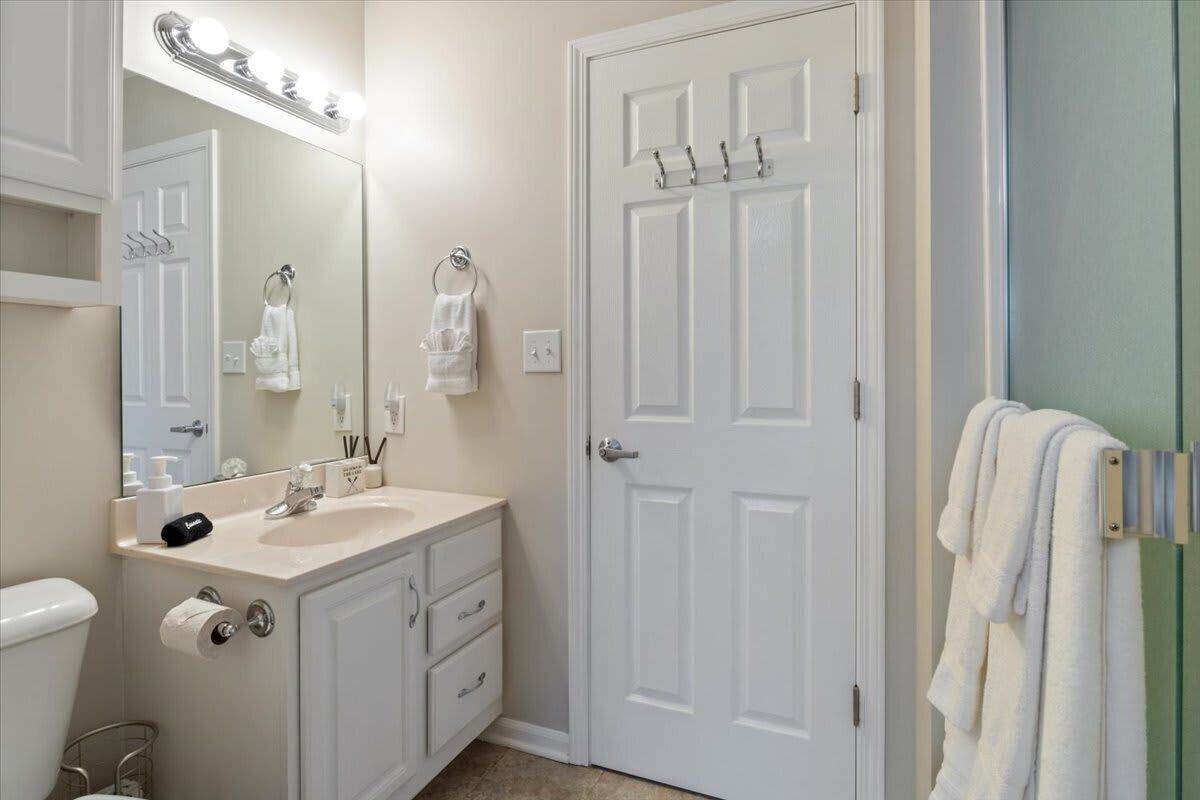 A bathroom with a white sink, mirror, and door. There are towels hanging on the door and a towel rack by the mirror.