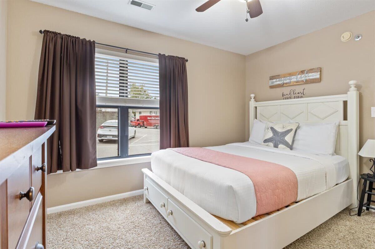 Une chambre à coucher avec un lit, une fenêtre avec des rideaux, un ventilateur de plafond et un mur orné d’une enseigne.
