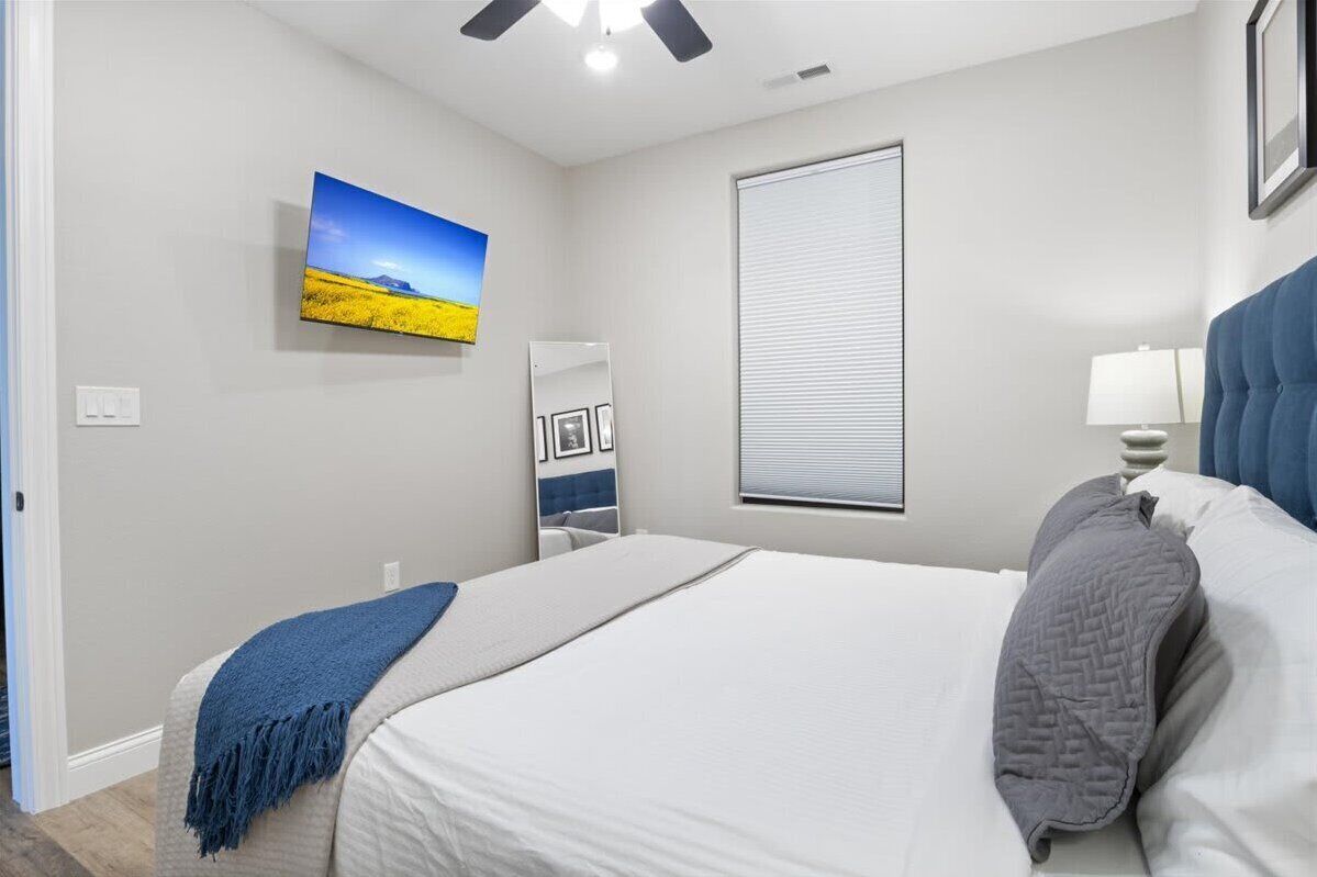 A bedroom with a bed, a ceiling fan, a window with blinds, a wall-mounted TV, and a bedside table with a lamp.