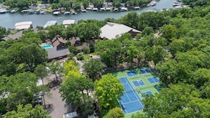 An aerial view of a lakeside property with a tennis court, boats docked along the shore, and dense forest surrounding the area. - Lakefront Condo Indoor Pool, Sauna, & Pickle Ball! (Osage Beach)