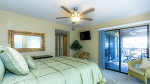 Un dormitorio con una cama grande, ventilador de techo, televisor y puerta corrediza de vidrio que da a un área exterior con mobiliario.