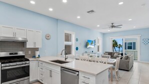 A modern kitchen with white cabinets, a stainless steel stove, and a dishwasher. There is a dining area with a white table and chairs, and a living room with a gray sofa and a TV. - 4BR Beachfront Balcony Condo | Partial Ocean View (Indian Shores)
