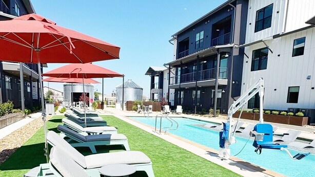 A pool area with lounge chairs and umbrellas, a clear sky, and modern buildings in the background. - The Nest @ Dovetail Cozy Condo in the Hill Country (New Braunfels)