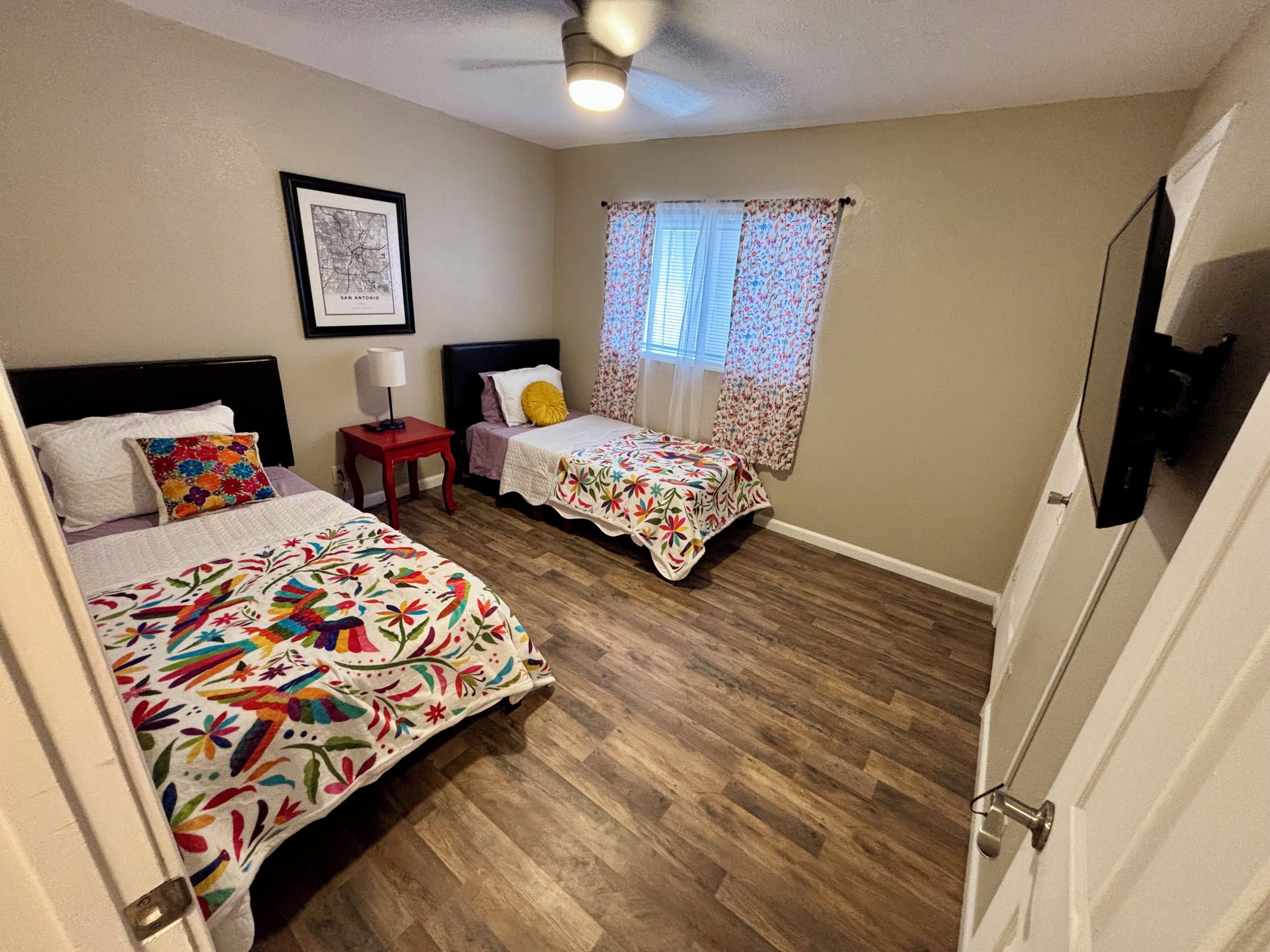 Une chambre avec deux lits, un ventilateur de plafond, une fenêtre avec des rideaux à motifs floraux et une carte encadrée accrochée au mur.