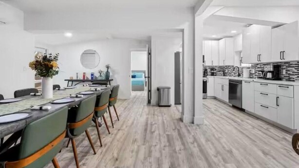 A modern kitchen with white cabinets, a dining area with a long table, and a view of the sea through an open door.