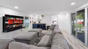 A modern living room with a large flat-screen TV, a grey sofa, and a glass door leading to an outdoor area.