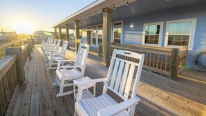 Een houten terras met witte Adirondack-stoelen, een overdekt gebied met lichtslingers en een gebouw met grote ramen.