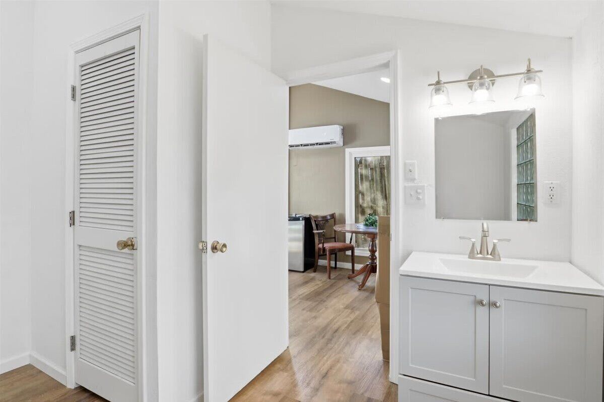 A room with a white door, a bathroom with a sink and mirror, and a dining area with chairs and a table.