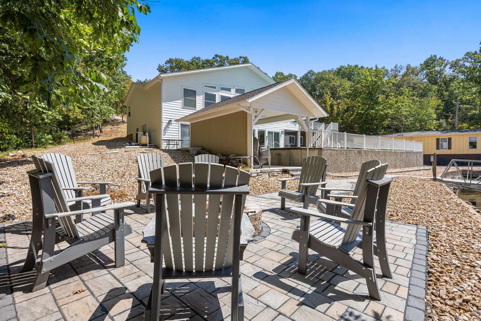A patio area with Adirondack chairs and a covered seating area, a white building with a covered porch, and a dock extending into a body of water.