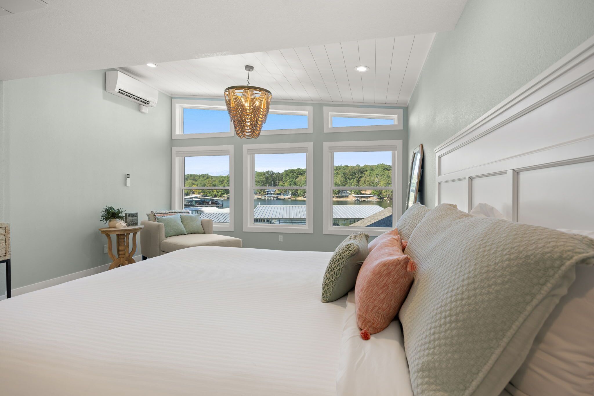Un dormitorio con una cama grande, varias ventanas, una lámpara colgante y vistas a un muelle y vegetación.