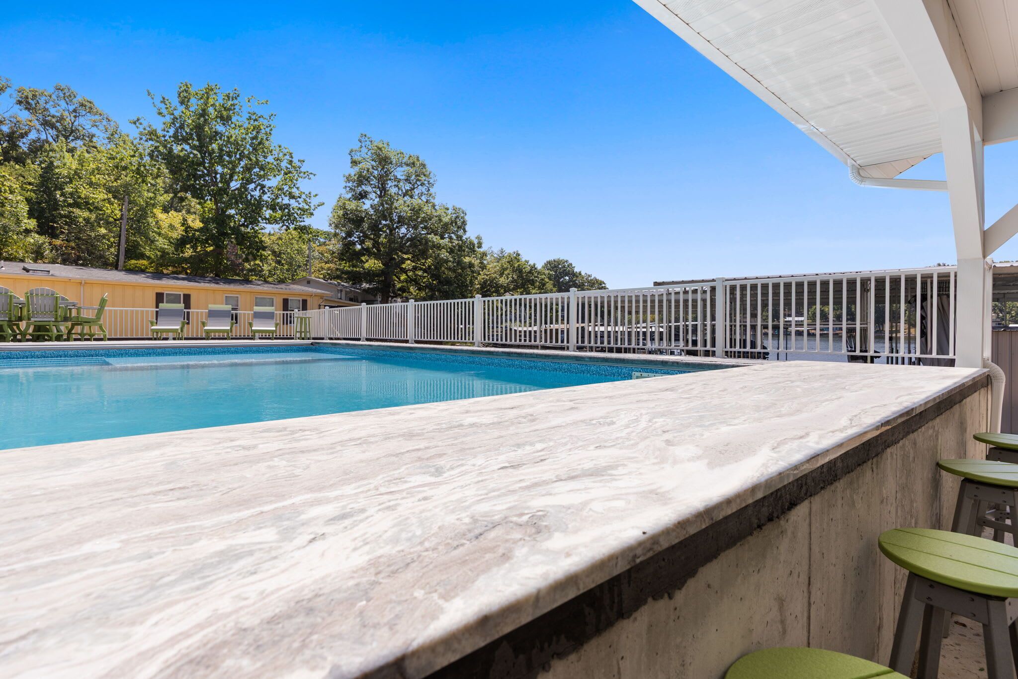 Piscina com deck branco e banquetas de bar verdes, cercada por uma cerca branca e árvores ao fundo.