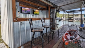 Zona de bar al aire libre con techo, paredes de metal, vigas de madera y dos televisores mostrando paisajes.