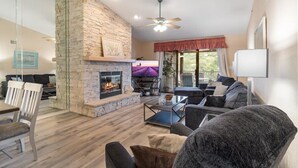 A living room with a stone fireplace, a ceiling fan, and a large window with a view of greenery. - Lakefront Condo Near the State Park (Osage Beach)
