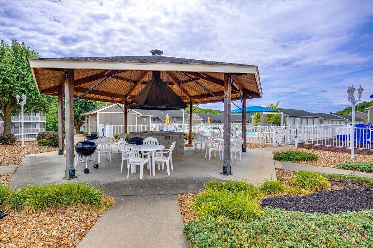 Ein Terrassenbereich im Freien mit überdachter Sitzgelegenheit, Grill und weißen Kunststoffstühlen.