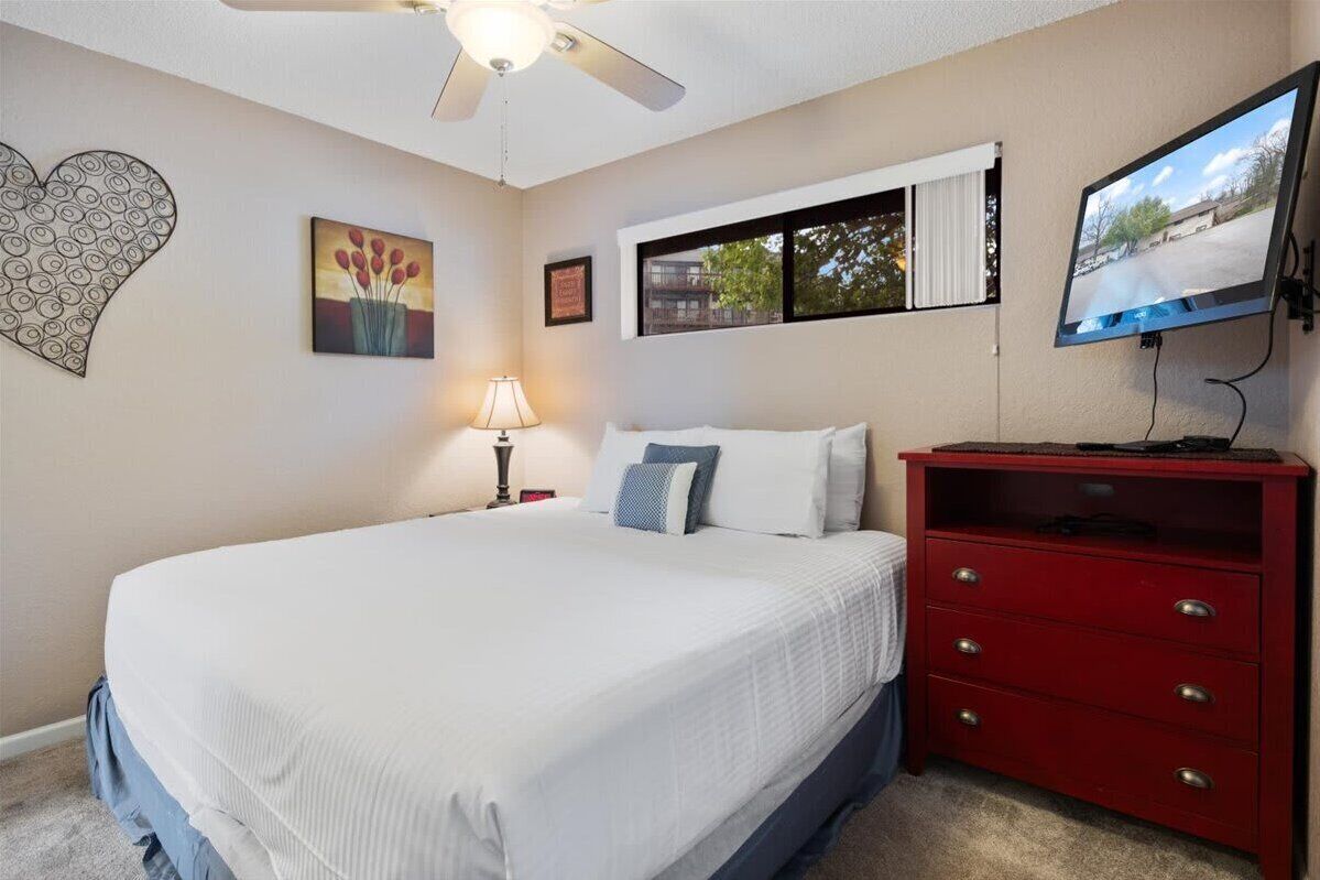 A bedroom with a bed, a red dresser, a wall-mounted TV, a ceiling fan, and artwork on the walls.