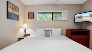 A hotel room with a bed, a nightstand with a lamp, a TV, a window with a view of trees, and a painting on the wall. - Lakefront Condo Near Margaritaville (Osage Beach)