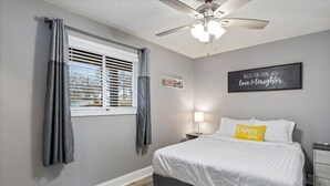 A bedroom with a bed, a ceiling fan, a window with curtains, a bedside table with a lamp, and motivational wall art. - Happy Harbor Lakefront Condo (Osage Beach)
