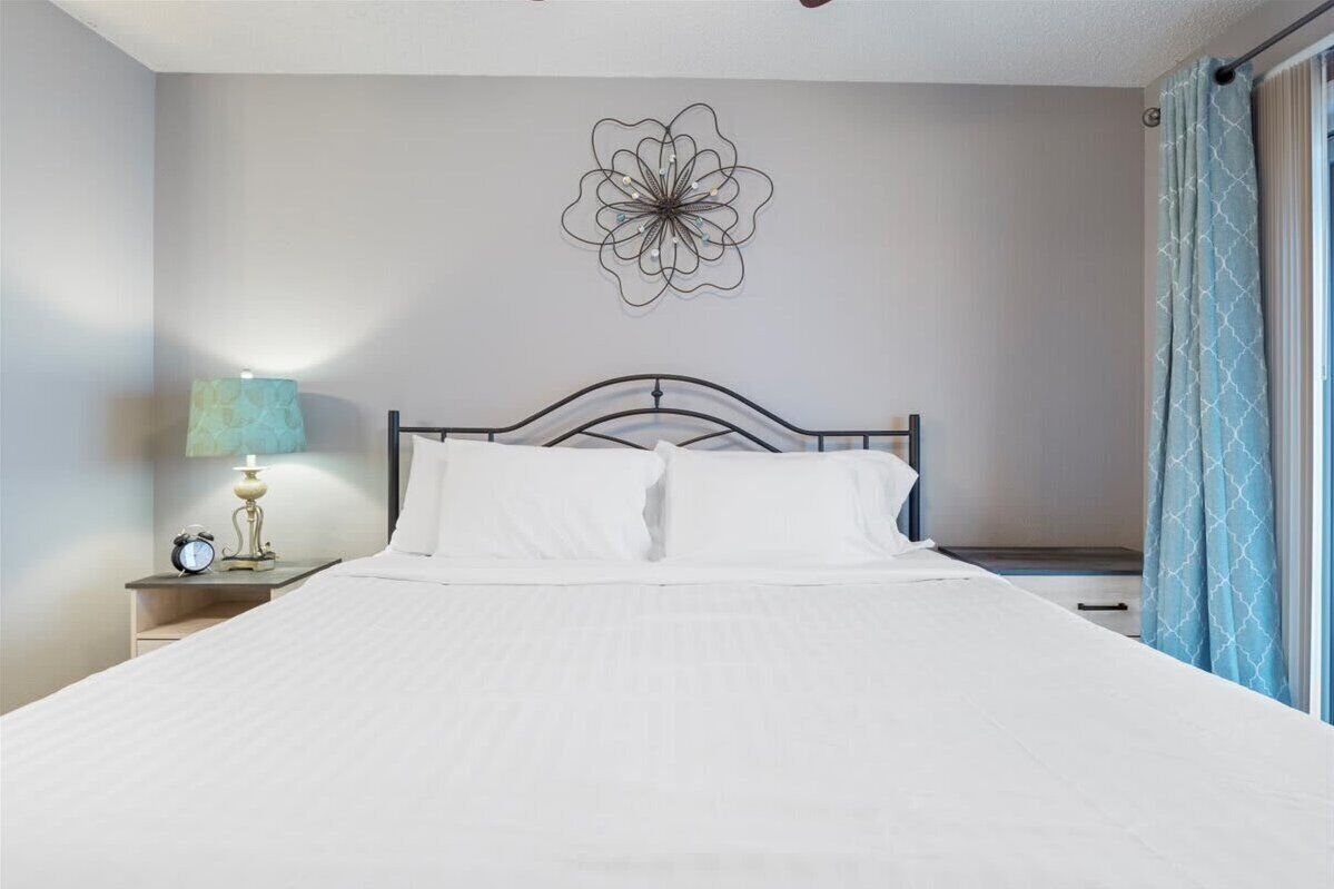 Una cama bien hecha con ropa de cama blanca, una decoración floral en la pared, una lámpara de noche y una ventana con cortinas.