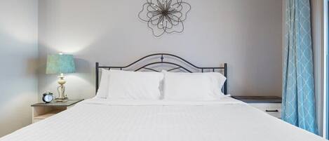 Una cama bien hecha con ropa de cama blanca, una decoración floral en la pared, una lámpara de noche y una ventana con cortinas.