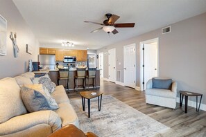 A spacious living room with a sofa, a coffee table, and a chair. There is a kitchen area with wooden cabinets and a dining area with chairs. - Happy Harbor Lakefront Condo (Osage Beach)