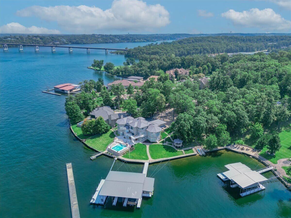 An aerial view of a lakeside property with a dock, a swimming pool, and a large house surrounded by trees.